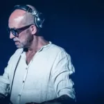 DJ Tomcraft se produisant sur scène, portant une chemise blanche, un casque et des lunettes, concentré sur son équipement.