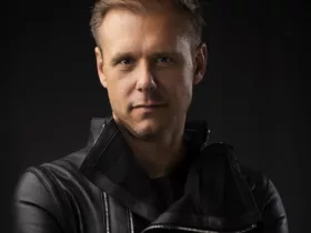 Portrait d'Armin van Buuren, bras croisés, vêtu d'une veste en cuir noir, avec un fond sombre.