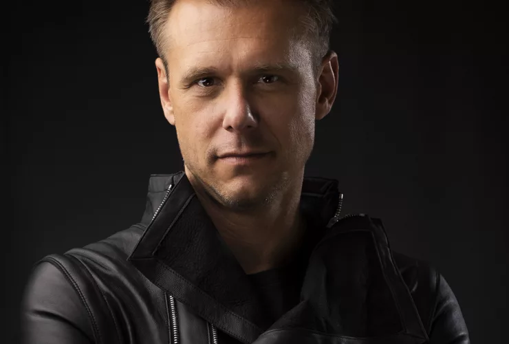 Portrait d'Armin van Buuren, bras croisés, vêtu d'une veste en cuir noir, avec un fond sombre.