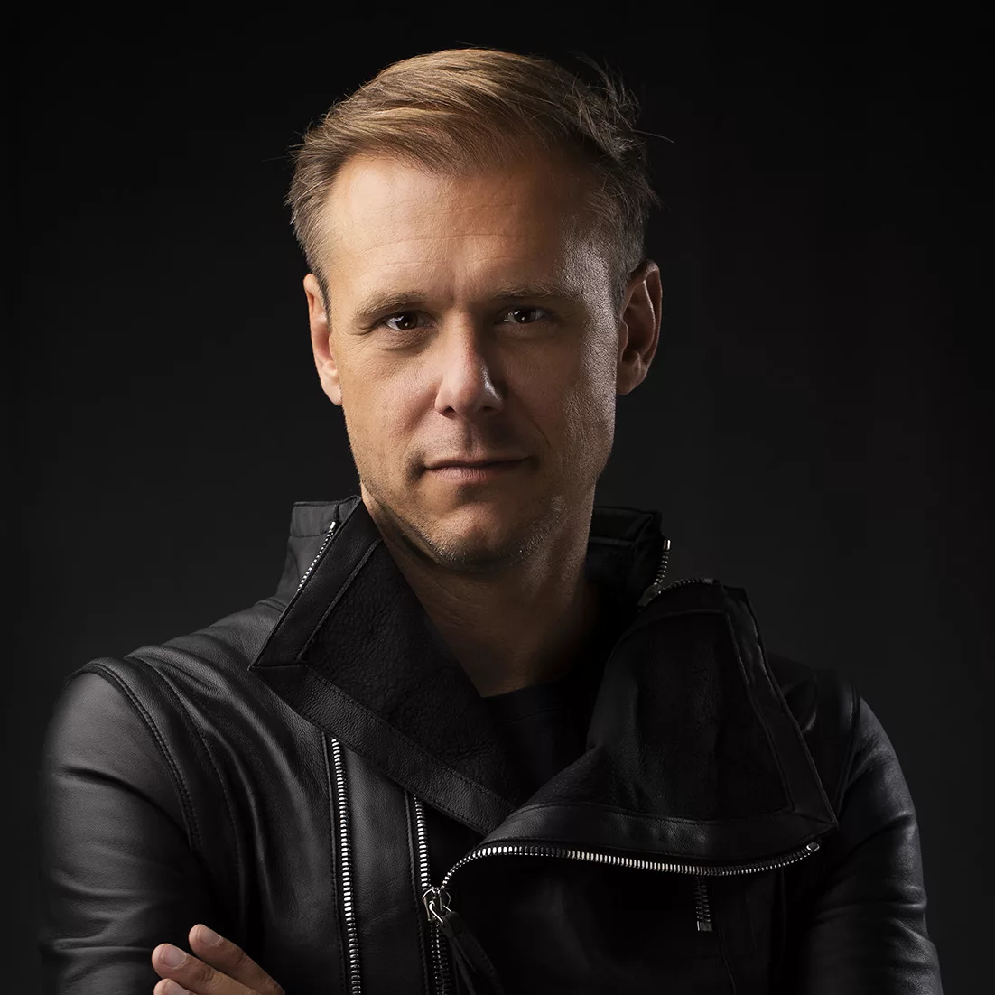 Portrait d'Armin van Buuren, bras croisés, vêtu d'une veste en cuir noir, avec un fond sombre.