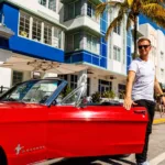 Armin van Buuren pose près d'une Ford Mustang rouge décapotable devant un hôtel art déco à Miami, en tenue décontractée avec un t-shirt blanc et des lunettes de soleil.