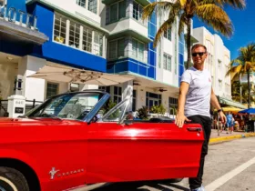 Armin van Buuren pose près d'une Ford Mustang rouge décapotable devant un hôtel art déco à Miami, en tenue décontractée avec un t-shirt blanc et des lunettes de soleil.