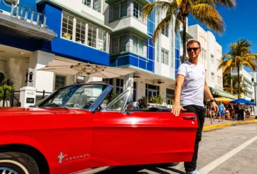 Armin van Buuren pose près d'une Ford Mustang rouge décapotable devant un hôtel art déco à Miami, en tenue décontractée avec un t-shirt blanc et des lunettes de soleil.