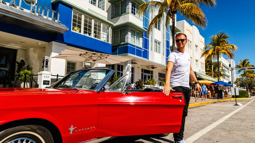 Armin van Buuren pose près d'une Ford Mustang rouge décapotable devant un hôtel art déco à Miami, en tenue décontractée avec un t-shirt blanc et des lunettes de soleil.