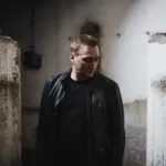 Paul van Dyk debout dans un espace industriel austère, vêtu d'un blouson en cuir noir et d'un t-shirt sombre, avec le regard baissé.