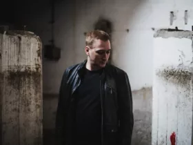 Paul van Dyk debout dans un espace industriel austère, vêtu d'un blouson en cuir noir et d'un t-shirt sombre, avec le regard baissé.