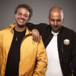 Les deux membres du duo de Psytrance Vini Vici posent ensemble. L'un porte une veste jaune et sourit, tandis que l'autre, en veste noire, a une expression plus posée.