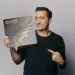 Paul van Dyk pose avec la réédition 30e anniversaire du classique Trance "For An Angel", affichant un sourire et pointant la pochette ornée d’une aile d'ange.