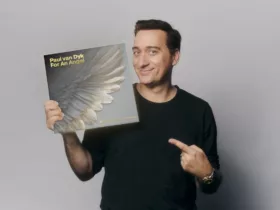 Paul van Dyk pose avec la réédition 30e anniversaire du classique Trance "For An Angel", affichant un sourire et pointant la pochette ornée d’une aile d'ange.