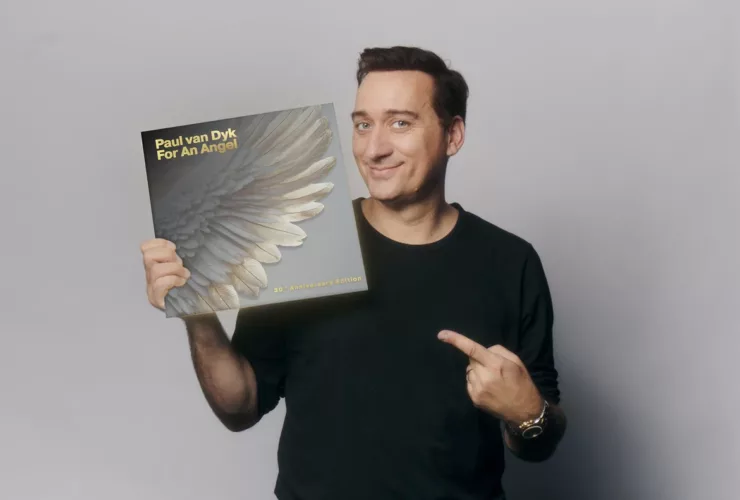 Paul van Dyk pose avec la réédition 30e anniversaire du classique Trance "For An Angel", affichant un sourire et pointant la pochette ornée d’une aile d'ange.