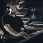 Ferry Corsten assis en studio, concentré devant son clavier, entouré de synthétiseurs et d’équipements de production musicale sur fond sombre.