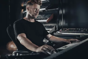 Ferry Corsten assis en studio, concentré devant son clavier, entouré de synthétiseurs et d’équipements de production musicale sur fond sombre.