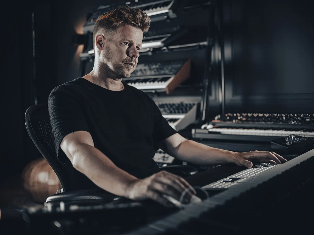 Ferry Corsten assis en studio, concentré devant son clavier, entouré de synthétiseurs et d’équipements de production musicale sur fond sombre.