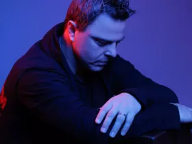 Markus Schulz, éclairé par une lumière bleue et rouge, pose pensivement dans un cadre minimaliste.