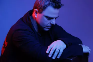 Markus Schulz, éclairé par une lumière bleue et rouge, pose pensivement dans un cadre minimaliste.