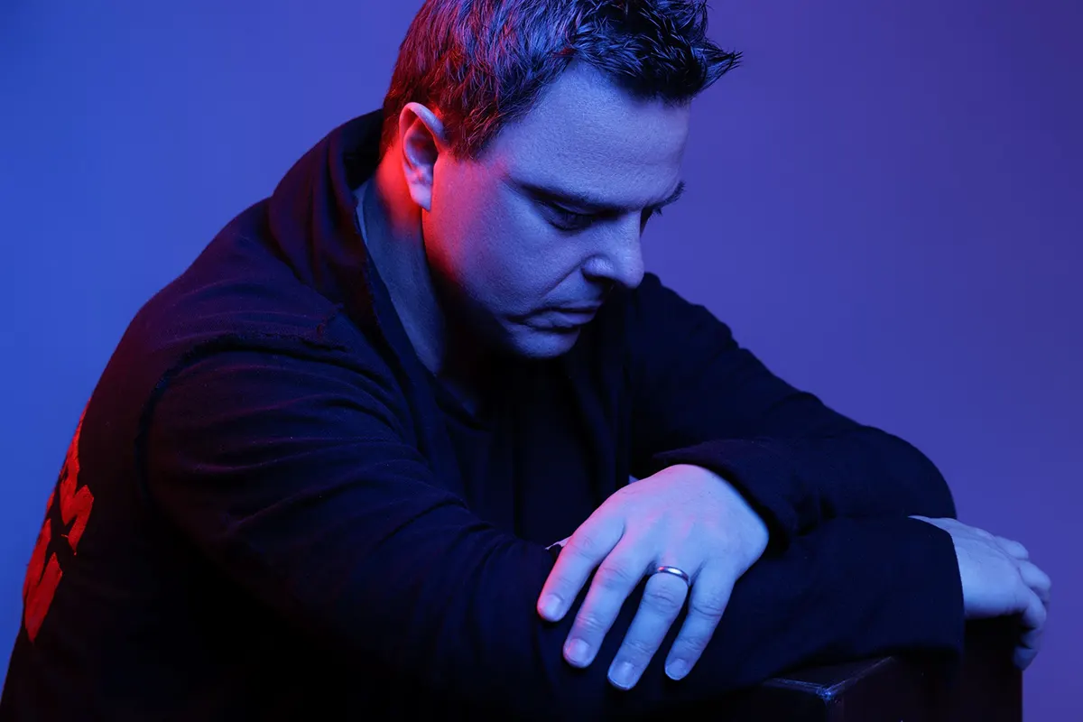 Markus Schulz, éclairé par une lumière bleue et rouge, pose pensivement dans un cadre minimaliste.