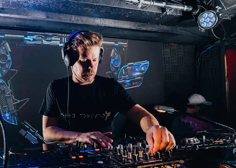 Ferry Corsten en pleine performance live, concentré derrière ses platines avec des visuels futuristes projetés en arrière-plan.