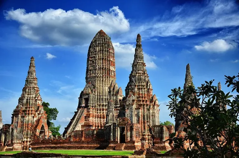 Vue du temple Wat Chaiwatthanaram à Ayutthaya, mettant en avant ses prangs majestueux en briques rouges sous un ciel bleu parsemé de nuages.