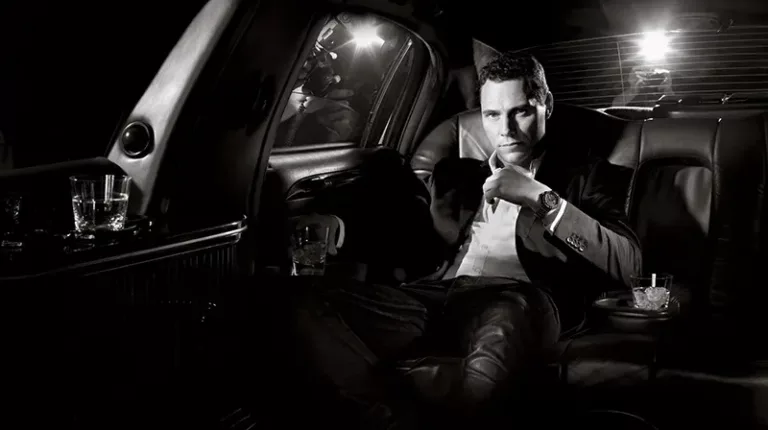 Tiësto dans une limousine, adoptant une attitude confiante et sophistiquée, élégamment vêtu et arborant une montre Guess luxueuse lors d'un shooting photo, avec un verre de whisky à la main, et des flashs illuminant l'arrière-plan.